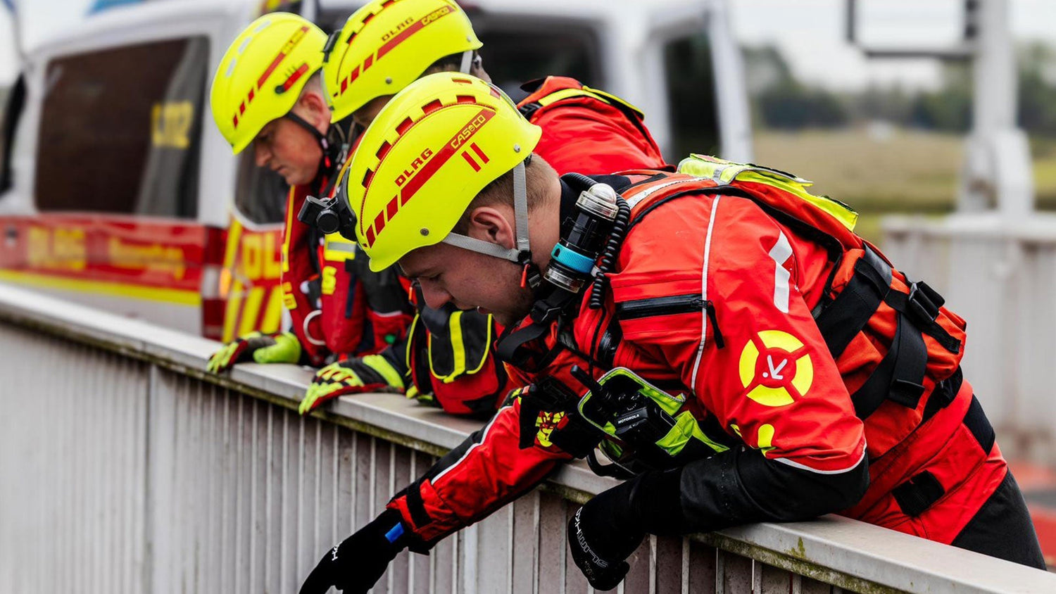 Rescue team in bright red water rescue suits and yellow helmets leaning over a bridge, preparing for a water rescue operation near an emergency vehicle.