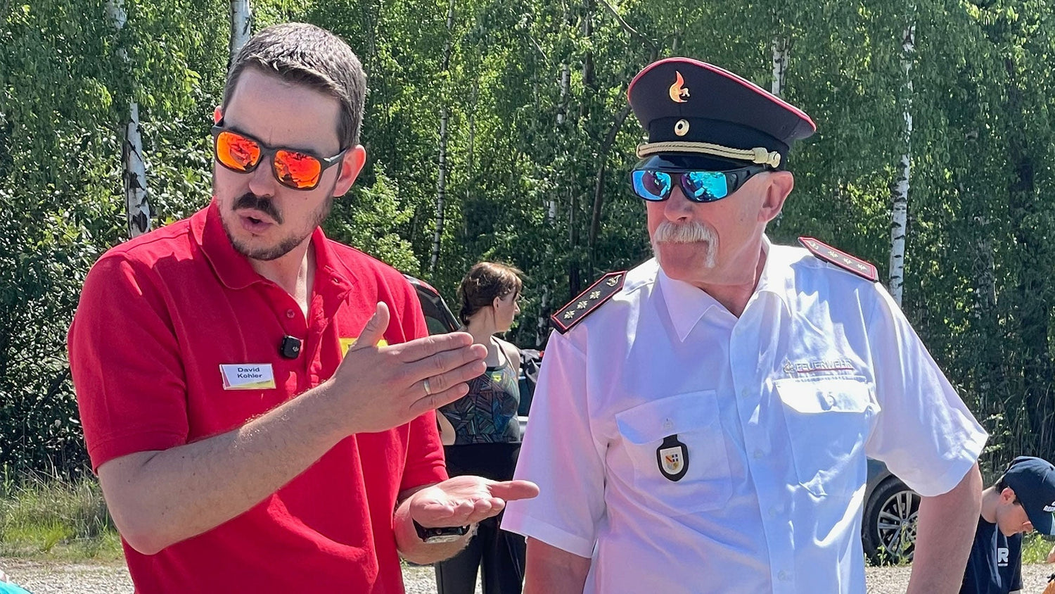 Two men in conversation outdoors at the RESTUBE Professional Experience Day 2026: David Kohler from RESTUBE in a red polo shirt gestures while talking with a fire brigade officer in white uniform and cap, surrounded by greenery and sunny weather