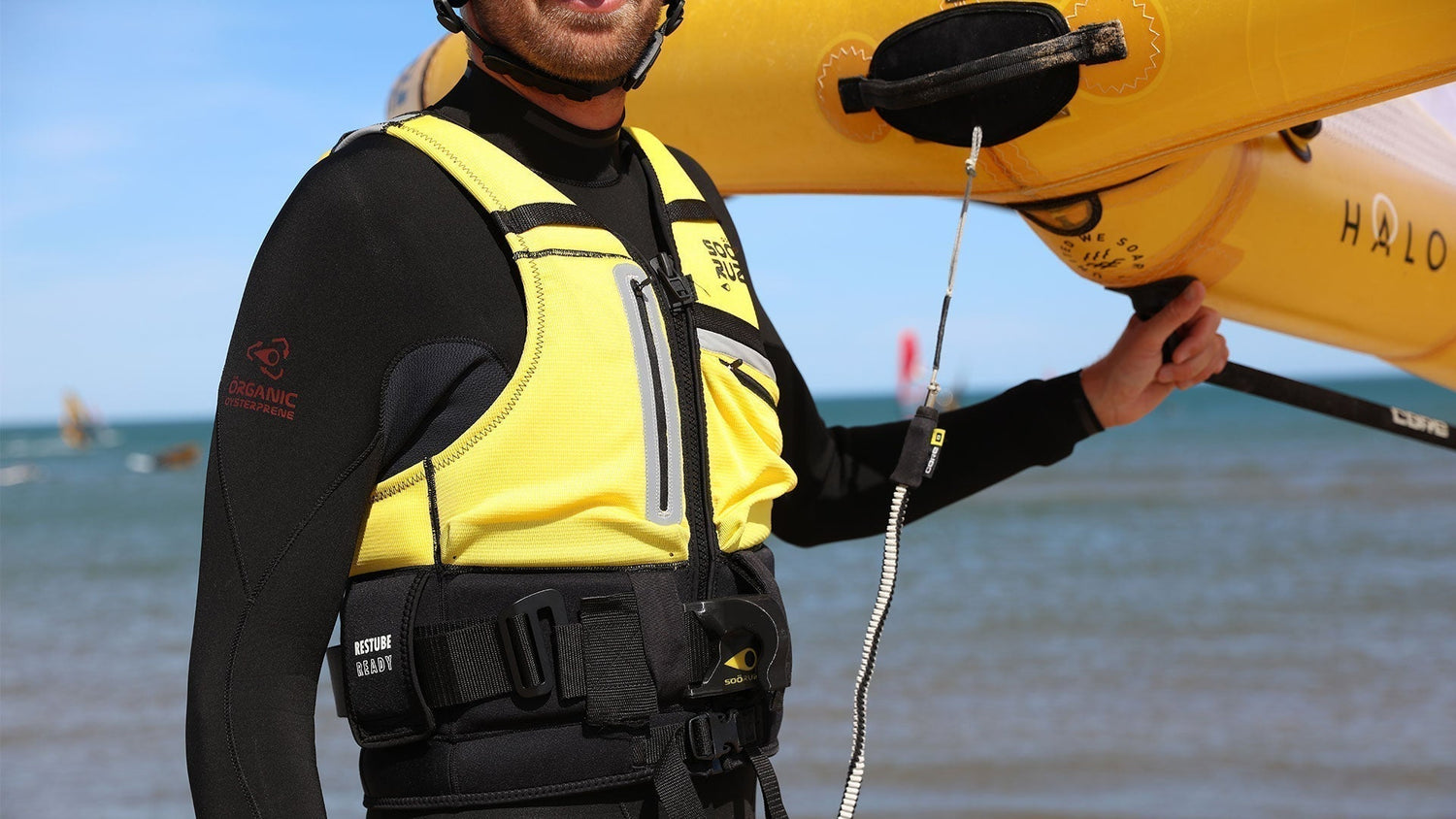 A person in a yellow impact vest and black wetsuit holding a yellow wing on a beach, wearing a harness with a “Restube Ready” attachment system.