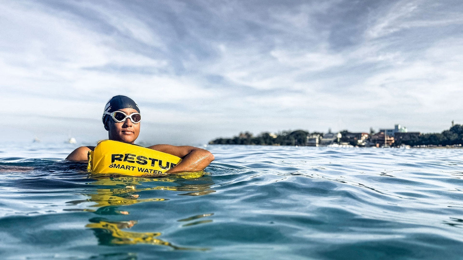 Female swimmer wearing a swim cap and goggles holding onto a yellow RESTUBE swim buoy in open water, coastline visible in the background