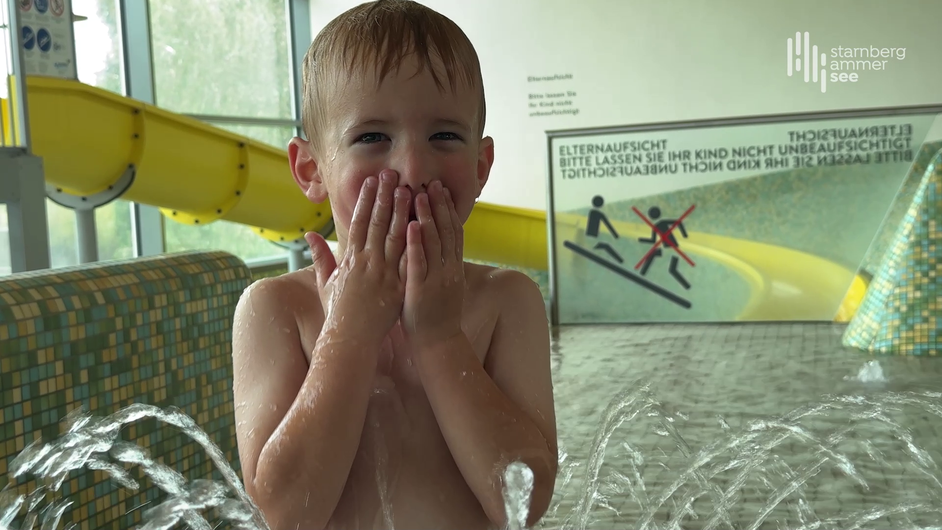 A smiling boy plays in the indoor area of a swimming pool in front of a yellow waterslide, with water fountains spraying from the floor – a joyful moment of water fun. Safety signage in the background highlights the importance of adult supervision.