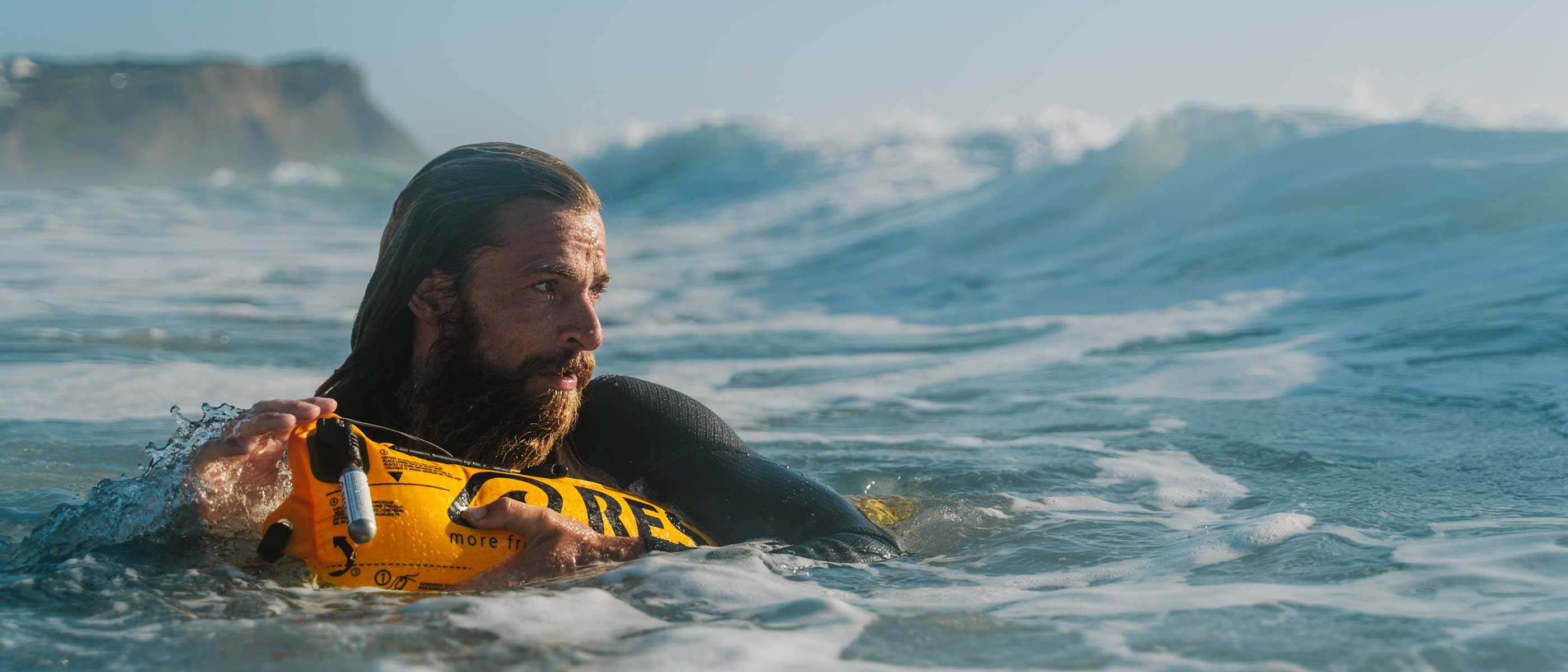 Surfer floating calmly with an inflated RESTUBE buoy near a rocky shore as a wave builds behind him.
