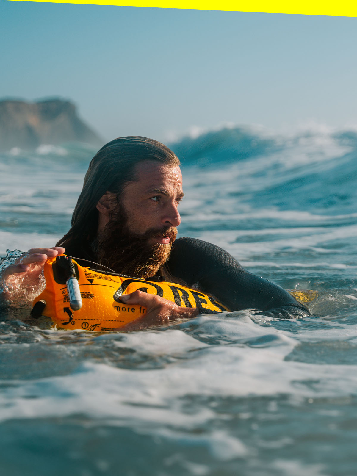 Man in the water with inflatable RESTUBE safety buoy for watersports, rough waves and cliffs on the background.
