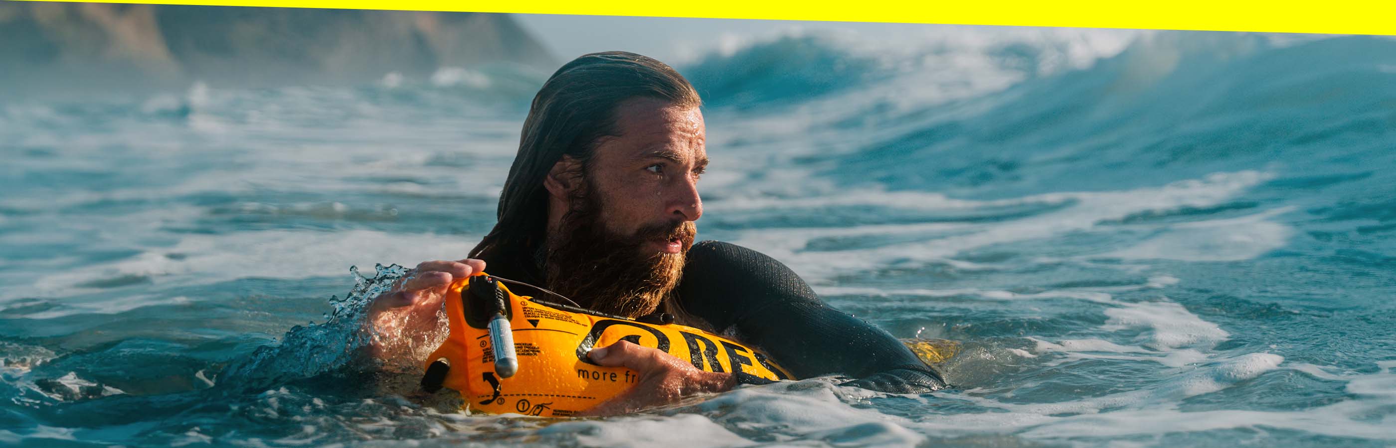 Surfer with long beard clinging calmly to an inflated yellow RESTUBE buoy in rough surf, waiting for the next wave.