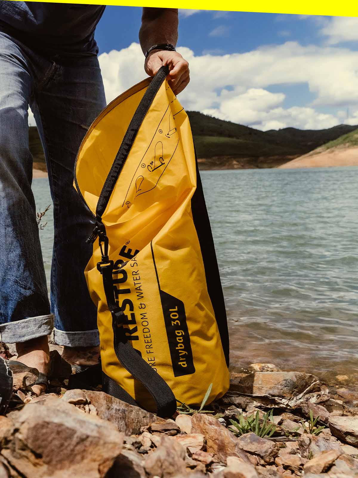 Person opening yellow RESTUBE Drybag 30L by the lake – for watersports activities.