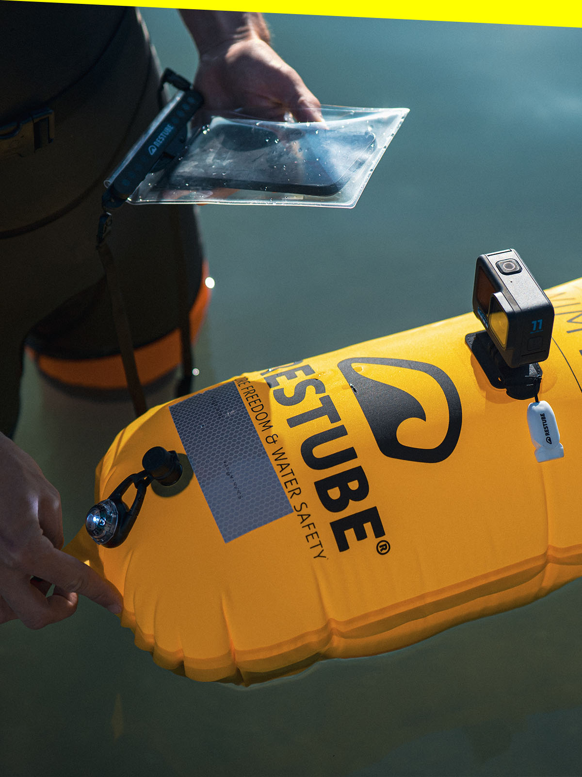 Detail shot: A yellow RESTUBE Swim Buoy equipped with action cam, signal light, and reflective patch; a watersports enthusiast holds a smartphone in a waterproof RESTUBE pouch.