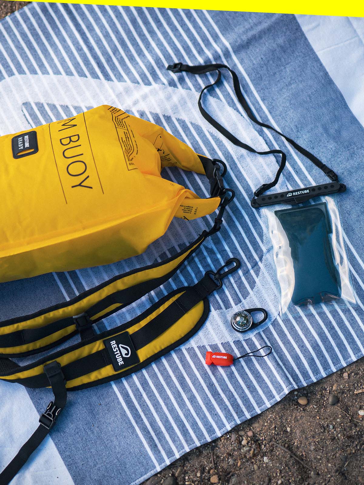 Close-up of a yellow RESTUBE Swim Buoy set on a striped towel – includes buoy, padded belt, waterproof phone pouch, mini compass, and signal whistle.