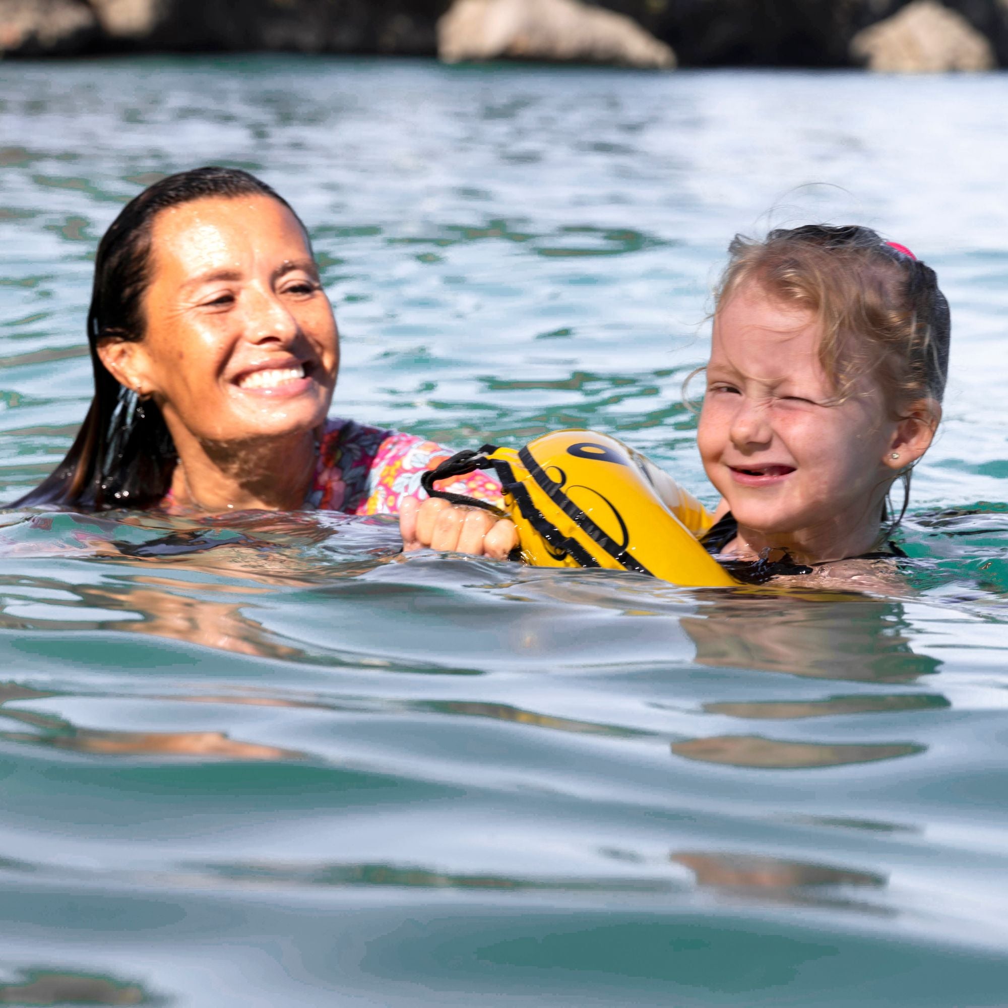 Smiling woman and young girl swimming in calm ocean water while holding a yellow swim buoy