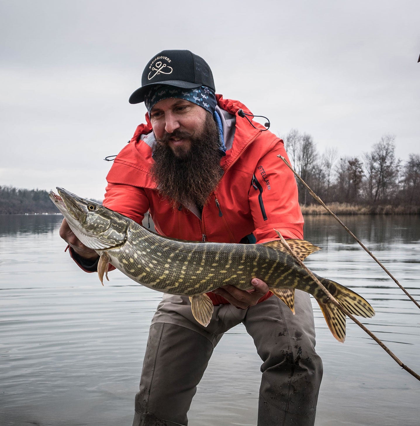 Bearded angler proudly holding a big pike at the lake’s edge – his red jacket and strong catch embody successful predator fishing.