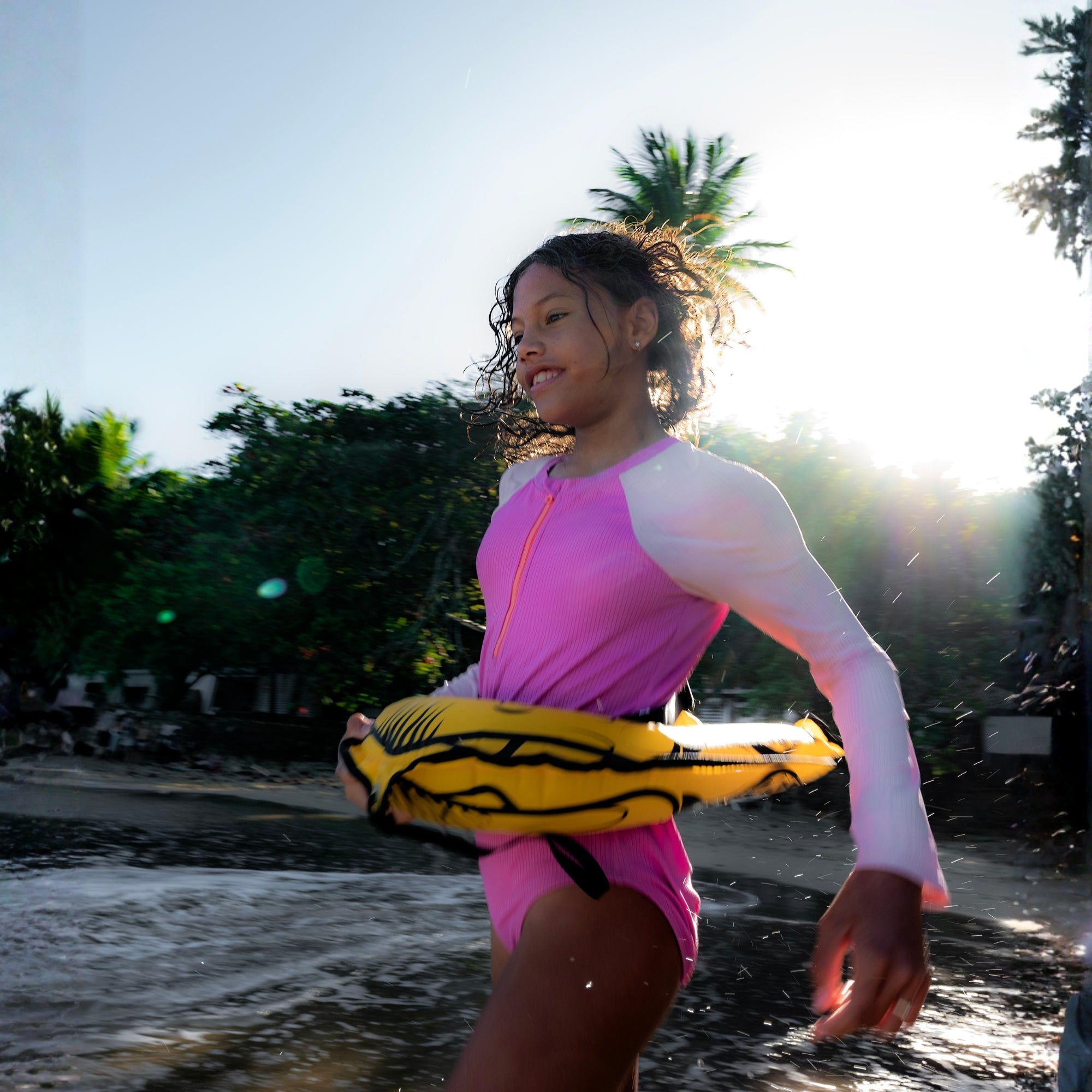 Girl running through beach waves holding RESTUBE JELLY inflatable water buddy, wearing pink swimsuit, sunny tropical setting