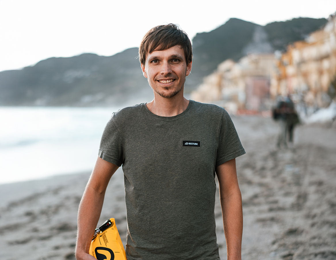 A man on the beach with RESTUBE safety buoy in hand and a RESTUBE T-shirt on - the CEO and Founder of RESTUBE – mountains and sea in the background.