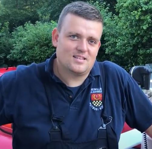 Young firefighter in navy polo with “Freiwillige Feuerwehr” crest, leaning casually on equipment – green background, helpful and focused.