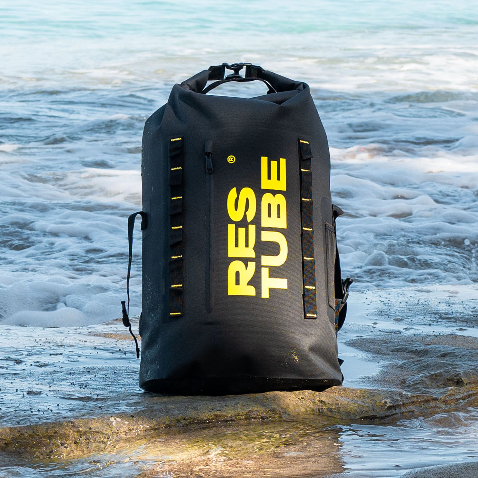 RESTUBE waterproof roll-top backpack standing on rocks at the shoreline, with waves in the background