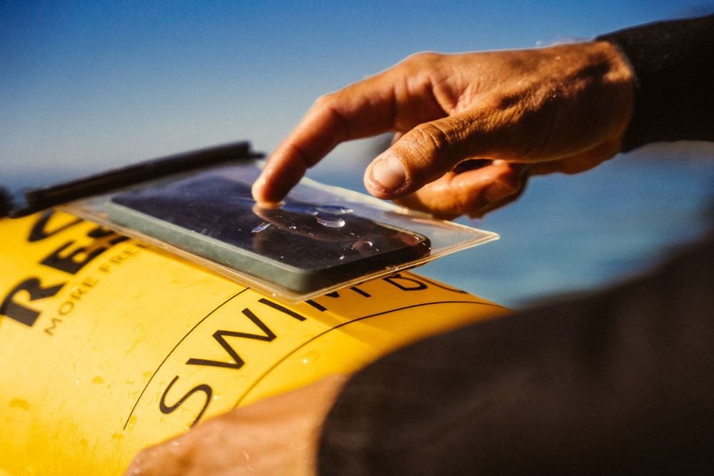 Waterproof phone case clipped onto a yellow RESTUBE buoy in action near water - a person tapping on the phone through the case.