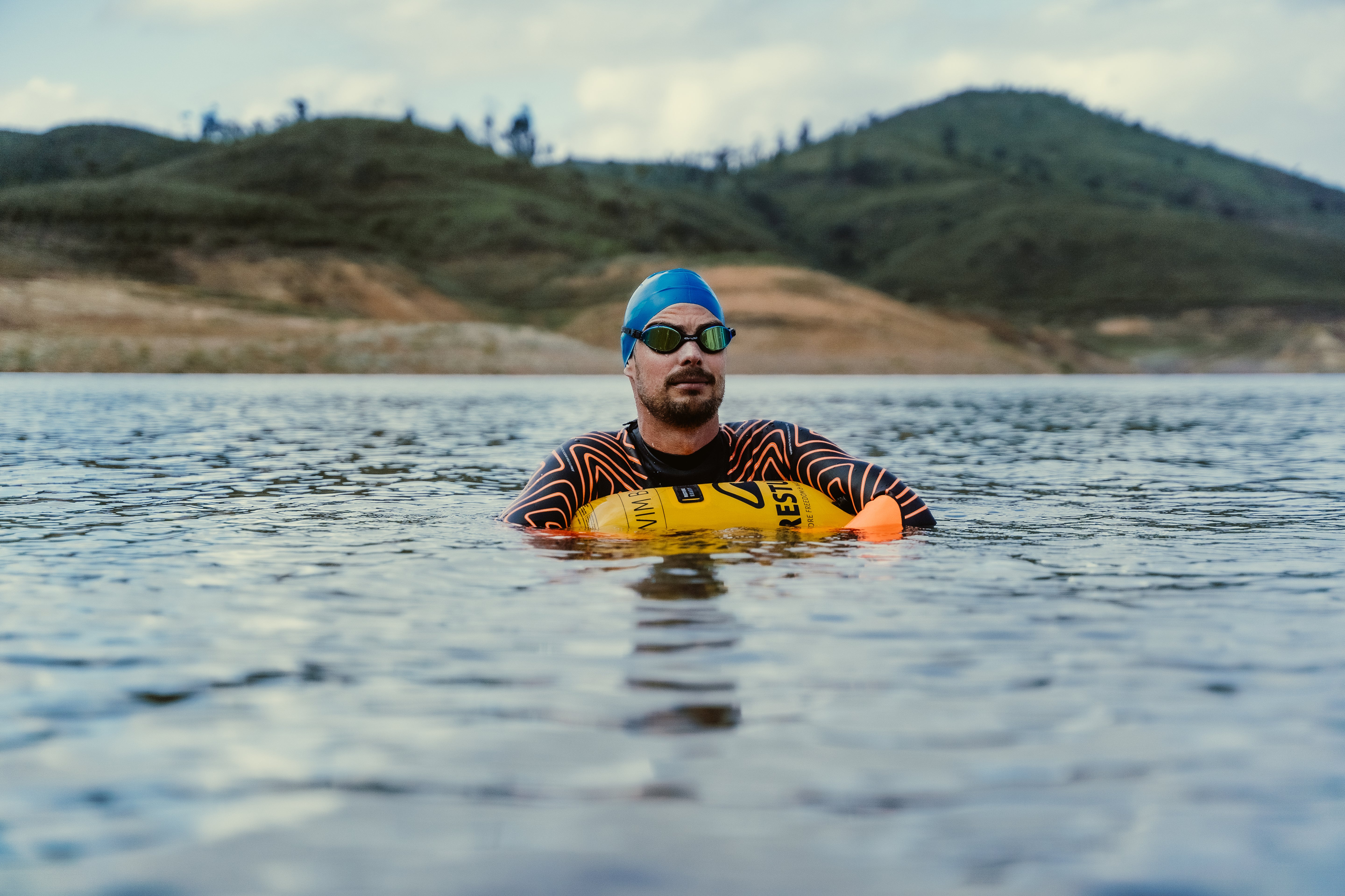 Swimmer with RESTUBE buoy in a lake, surrounded by hills – for water safety and visibility.