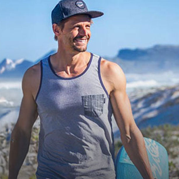 A man in a gray tank top and a gray cap with surfboard in mountain scenery.