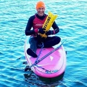 Person wearing a wetsuit and swim cap sitting on a pink stand-up paddleboard in calm blue water, holding a yellow rescue buoy.