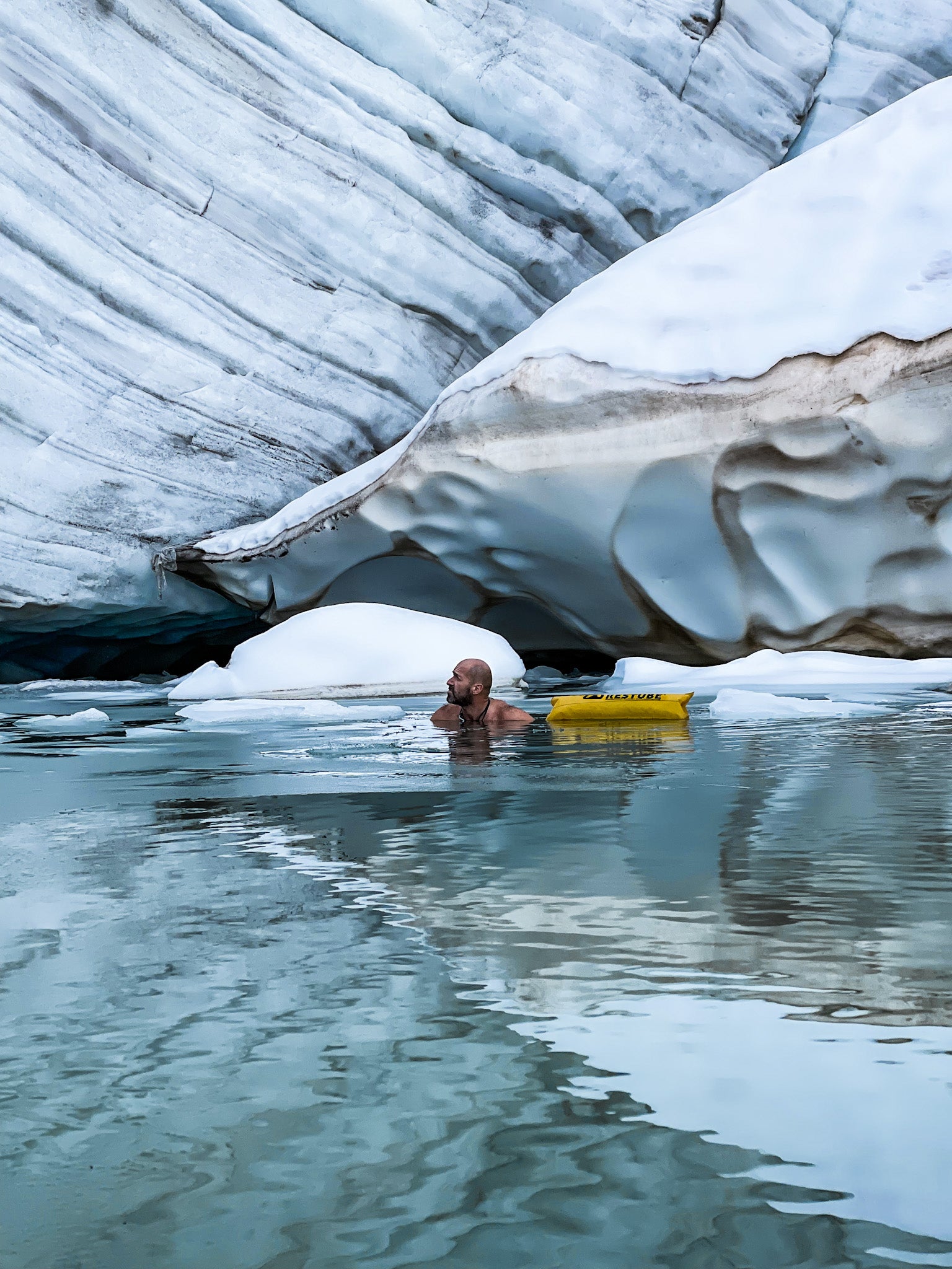 A man swimming in icy glacial water among ice floes, accompanied by a yellow RESTUBE buoy – safety even in extreme environments.