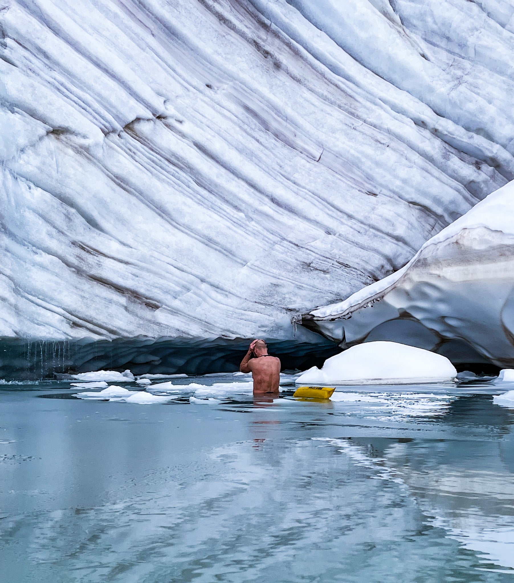 A man in icy water with yellow RESTUBE buoy in front of glacier wall.