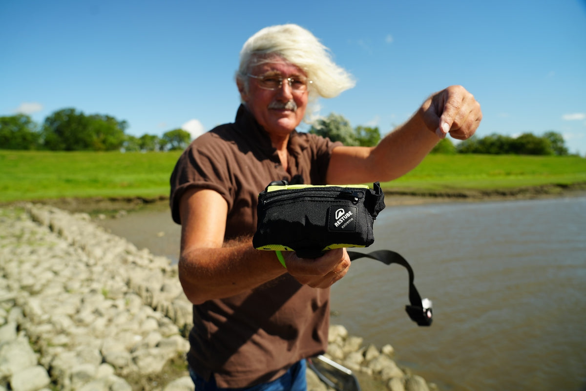 Jörg Ovens on a stone embankment by a tidal river, holding the RESTUBE EXTREME towards the camera and pointing at it
