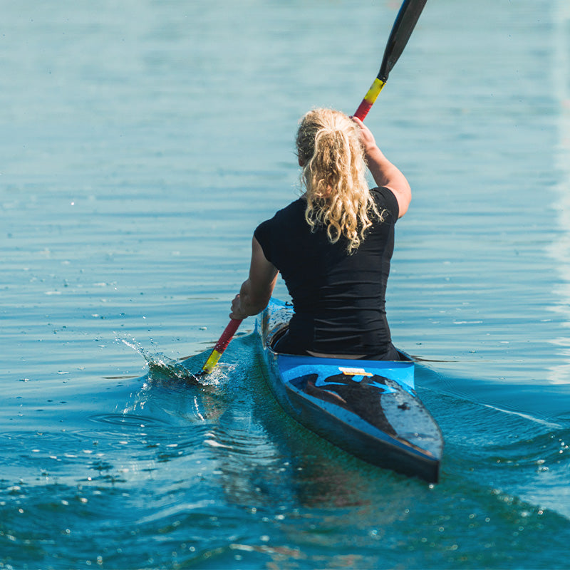 A kayaker on calm blue water, sitting with her back to the camera.