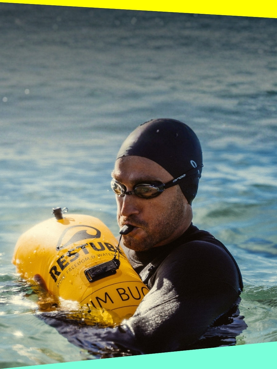 Open water swimmer blowing a whistle while holding a yellow RESTUBE Swim Buoy, wearing a wetsuit, cap and goggles
