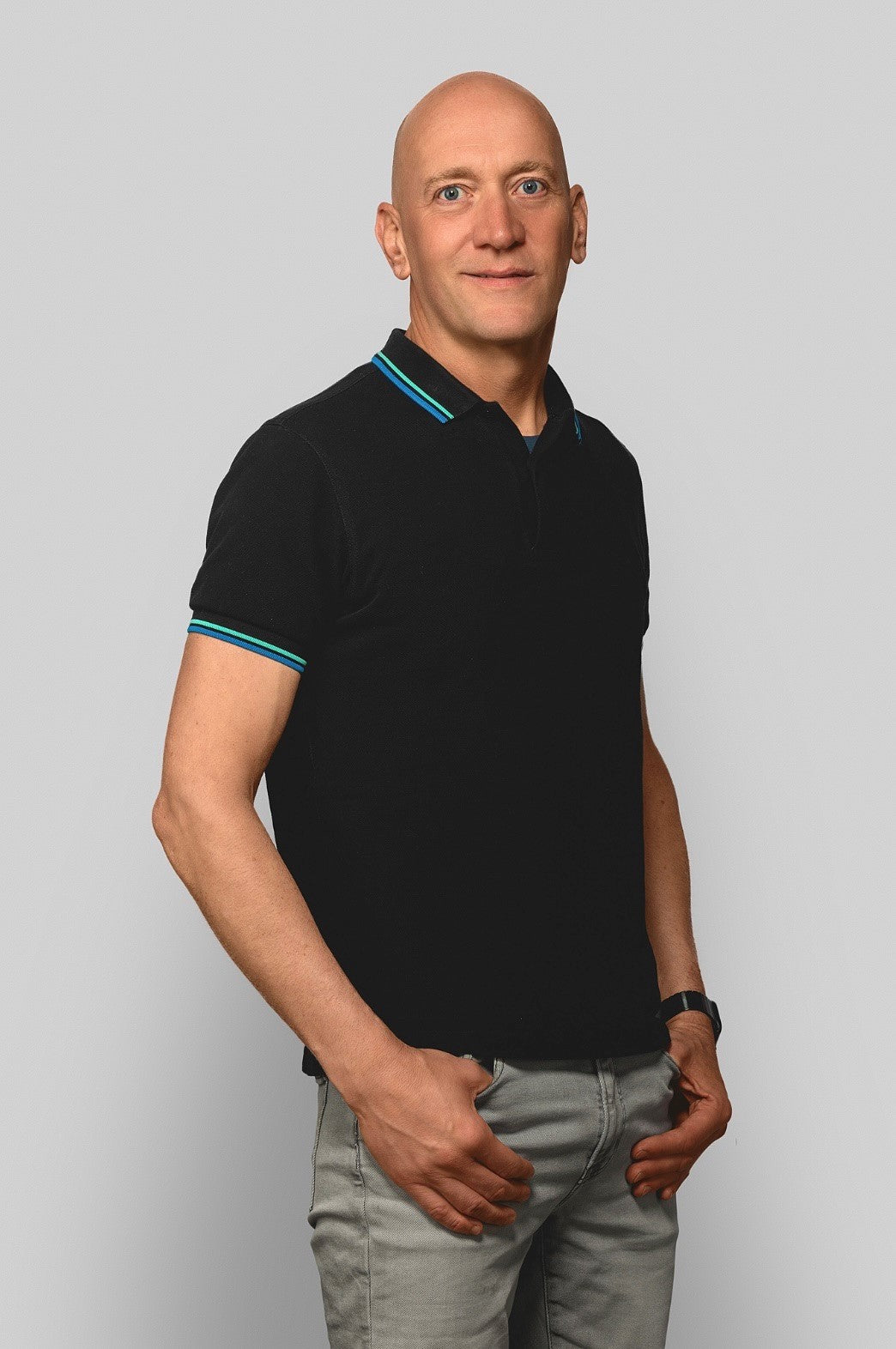 Full-body shot Bald man (Martin Fahnemann) in black polo with turquoise stripes and grey jeans – relaxed pose in grey studio setting.