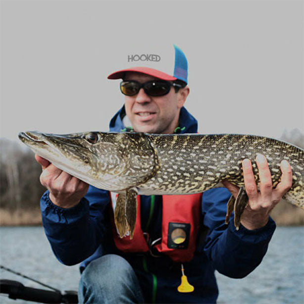 Happy angler wearing a "HOOKED" cap and sunglasses proudly shows off a large pike caught from a fishing boat – protected by a red life vest.