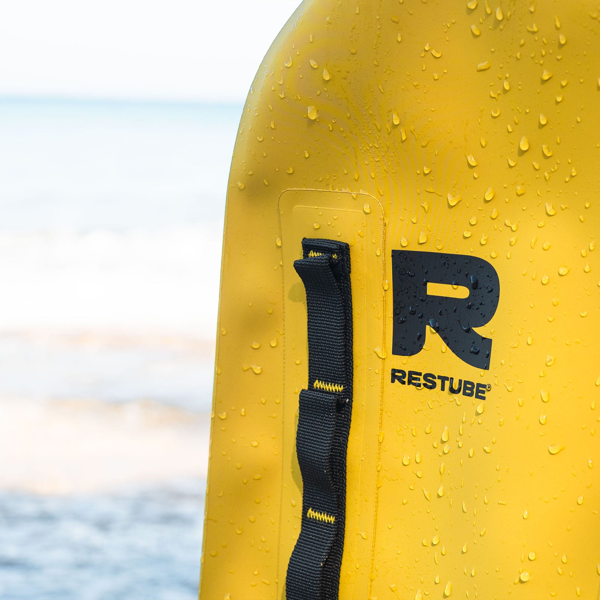 Close-up of a yellow RESTUBE waterproof backpack with water droplets, highlighting durable material and attachment loops by the sea