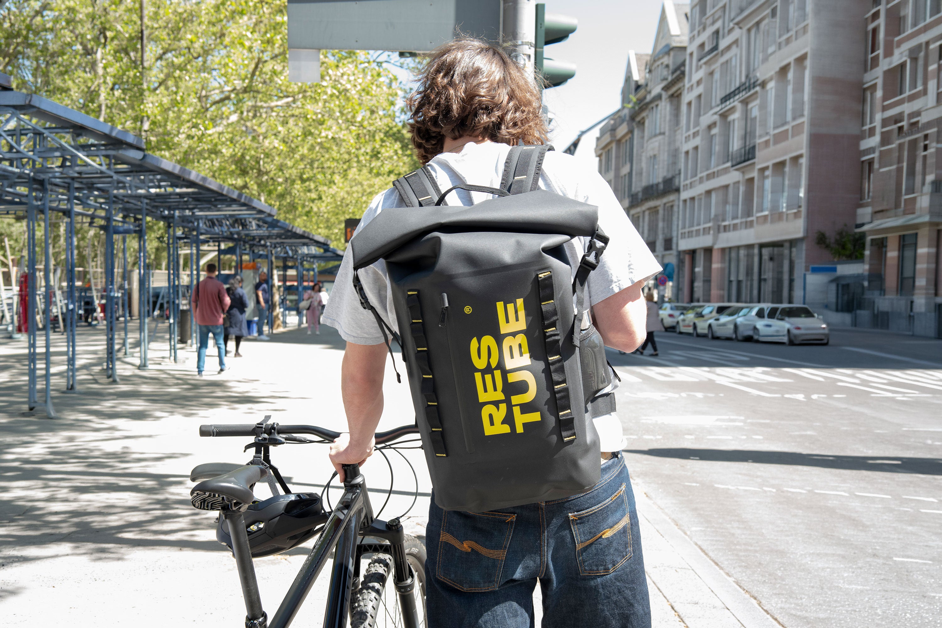 Man walking a bicycle through a sunny city street wearing a black RESTUBE COAST water-resistant backpack