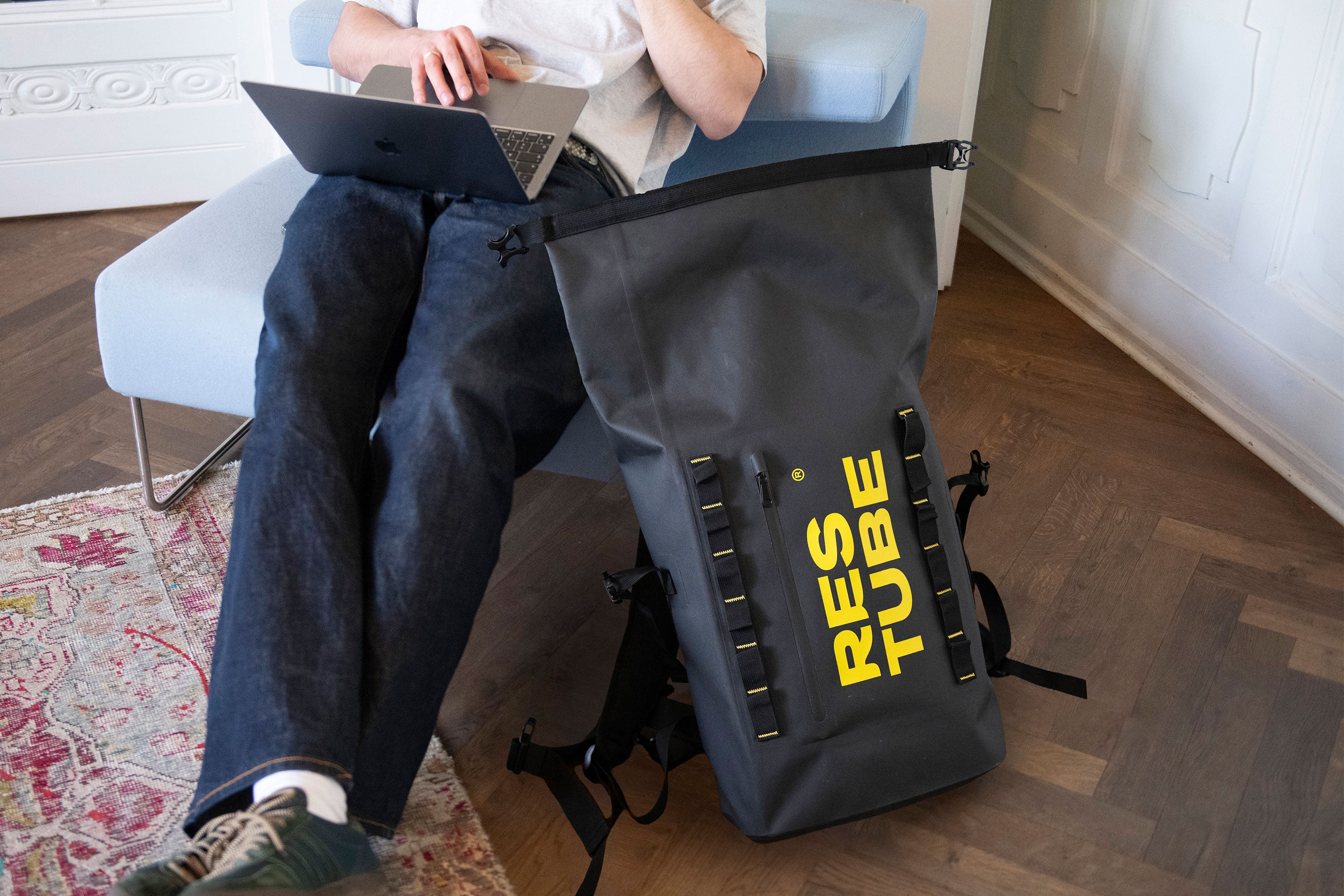 Person sitting in a chair working on a laptop with a black RESTUBE COAST water-resistant backpack placed on the floor beside the