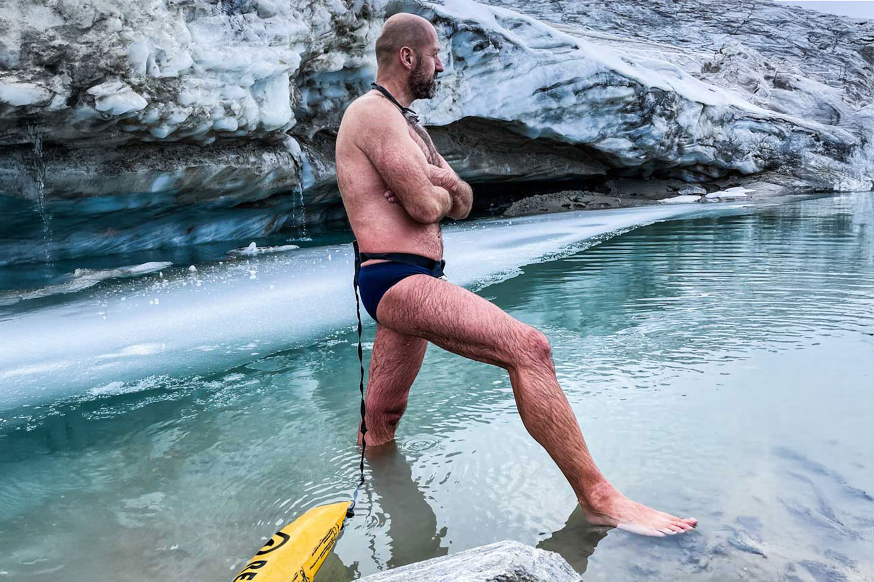 Man standing in icy glacier water wearing a RESTUBE safety buoy around his waist, preparing for a cold-water swim.