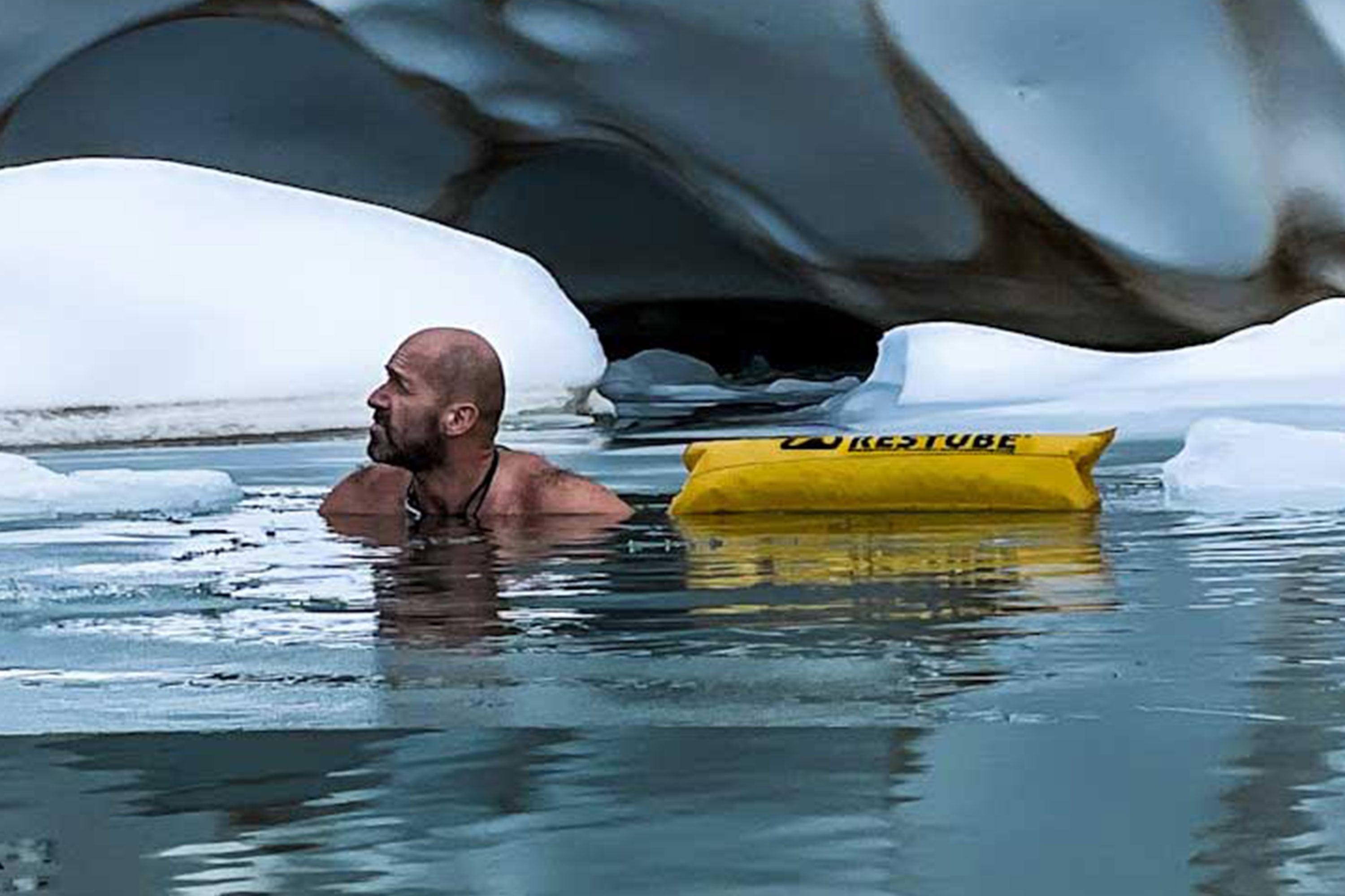 Man swimming in icy glacier water with an inflated yellow RESTUBE buoy floating beside him for safety.