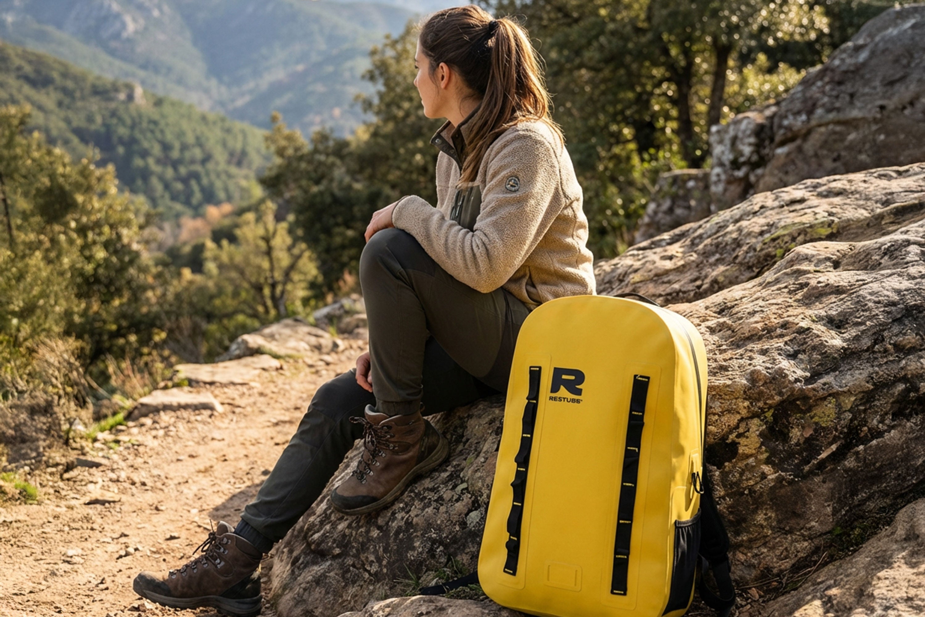 Woman sitting on rocks in a mountain landscape with a yellow waterproof backpack beside her.