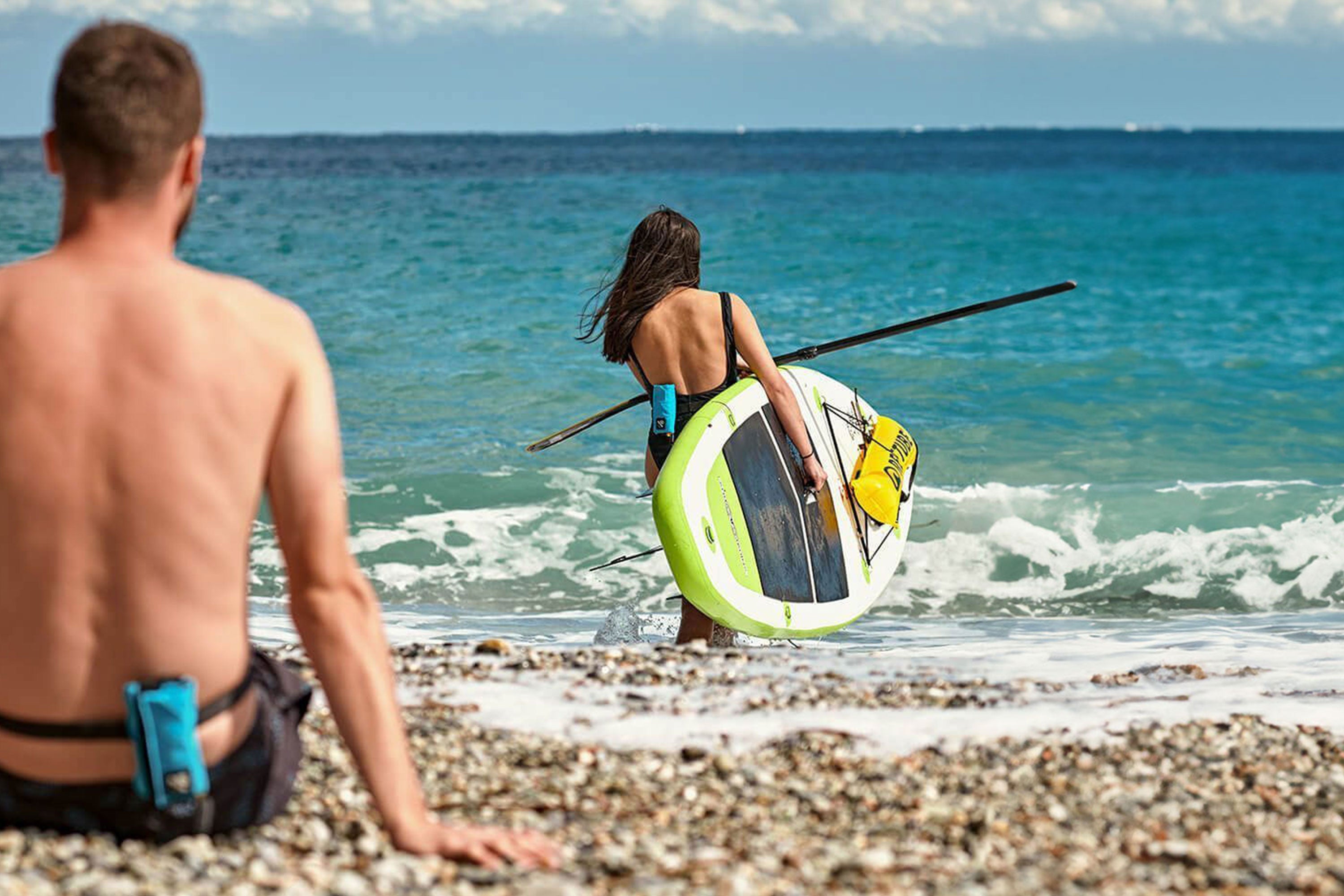 Woman with SUP board and RESTUBE buoy on the beach, carrying the board into the water, man with RESTUBE buoy in the foreground, sitting on the ground.