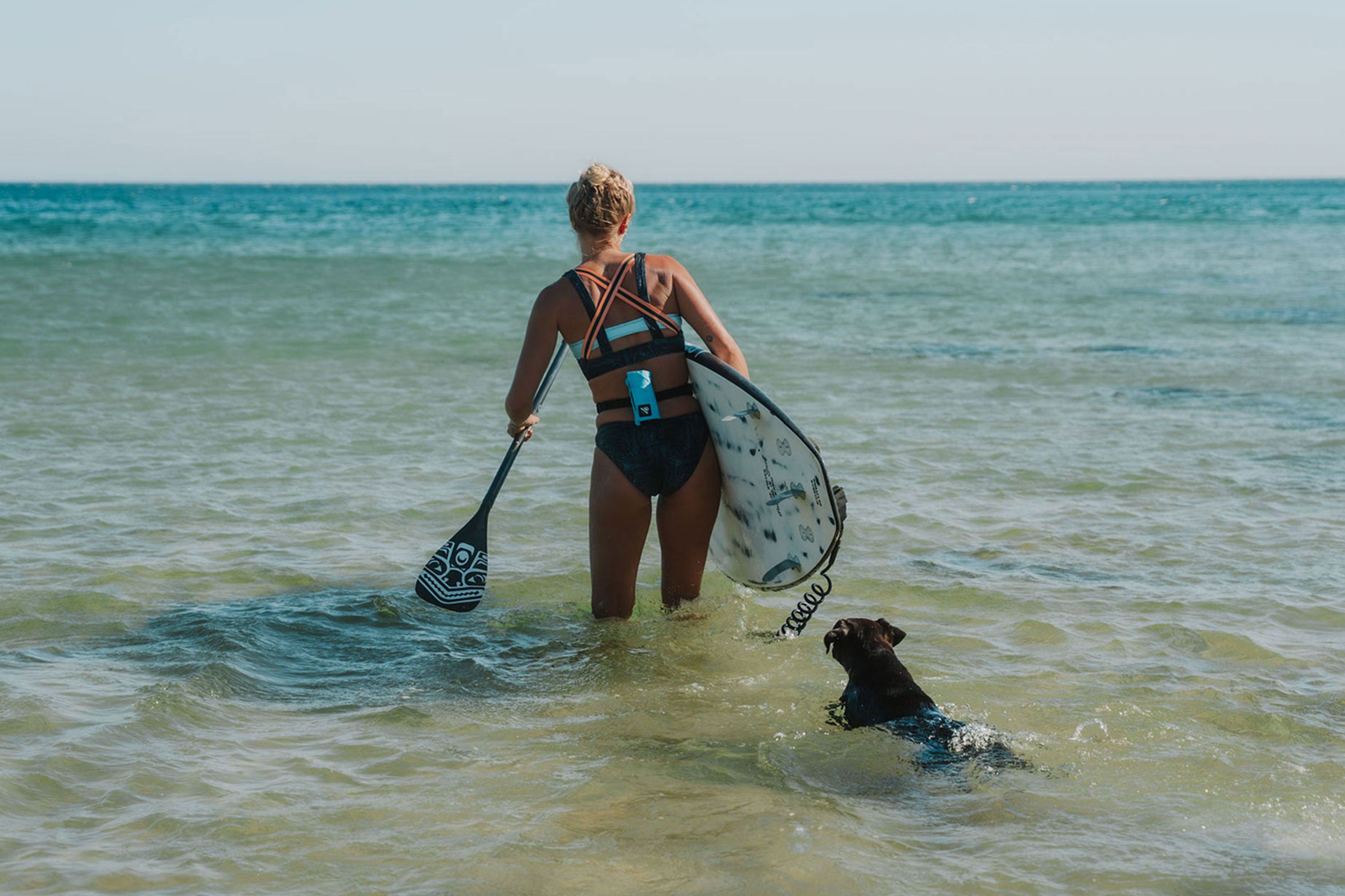 Female watersports athlete with RESTUBE buoy, carrying a board into the water, and a small dog swimming behind her in the ocean.