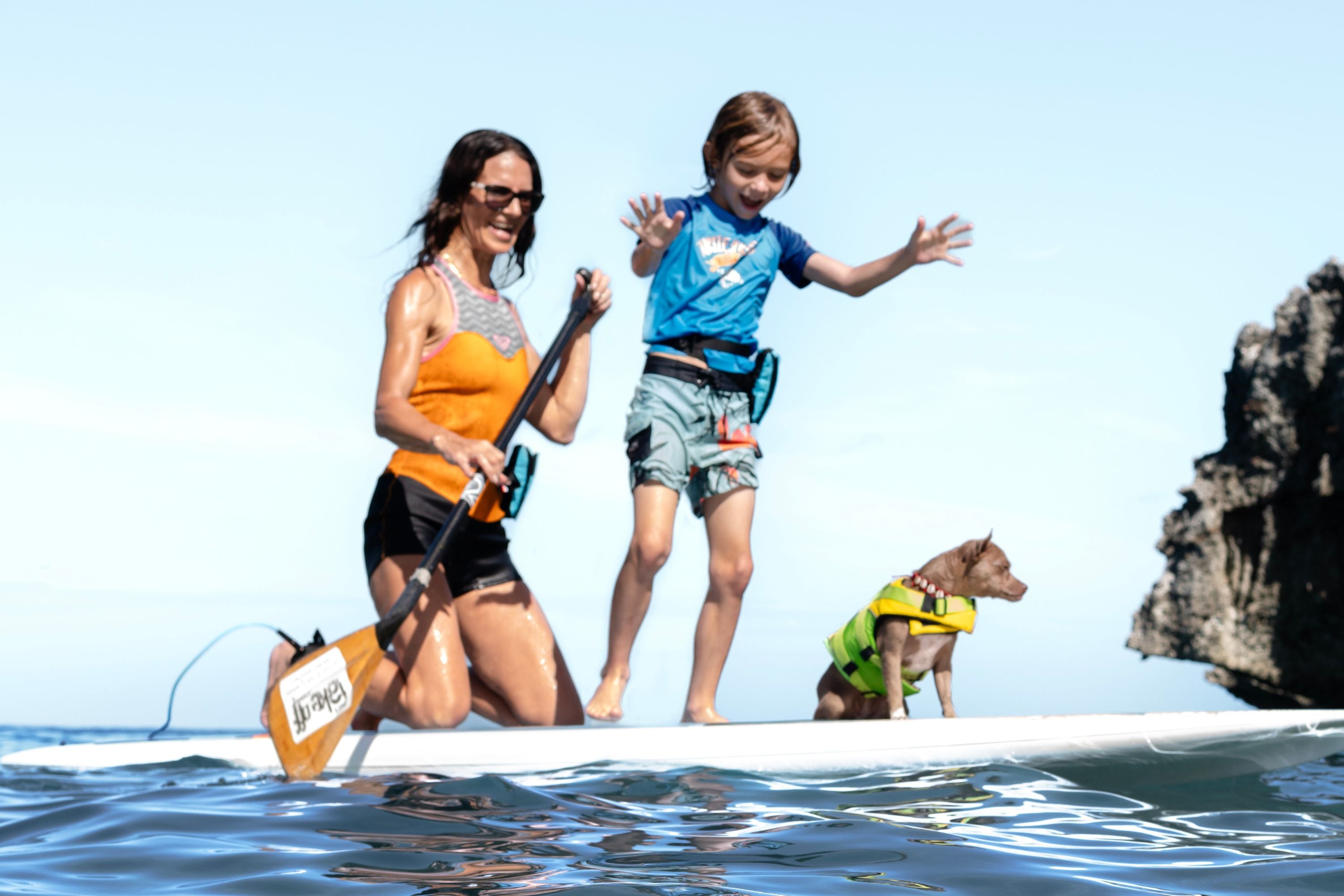 Woman and child standing on a paddleboard with a dog wearing a RESTUBE life jacket, near ocean rocks