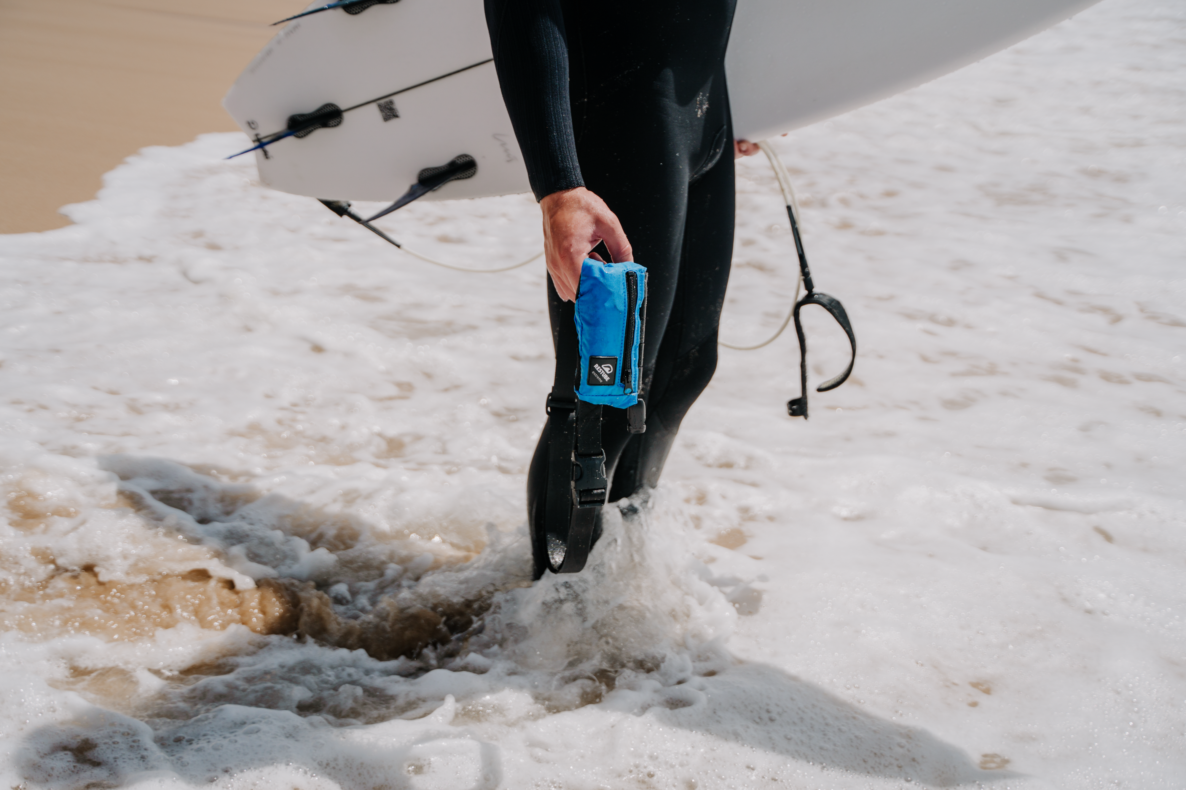 A surfer with RESTUBE buoy on his leg standing on the beach with a surf board.