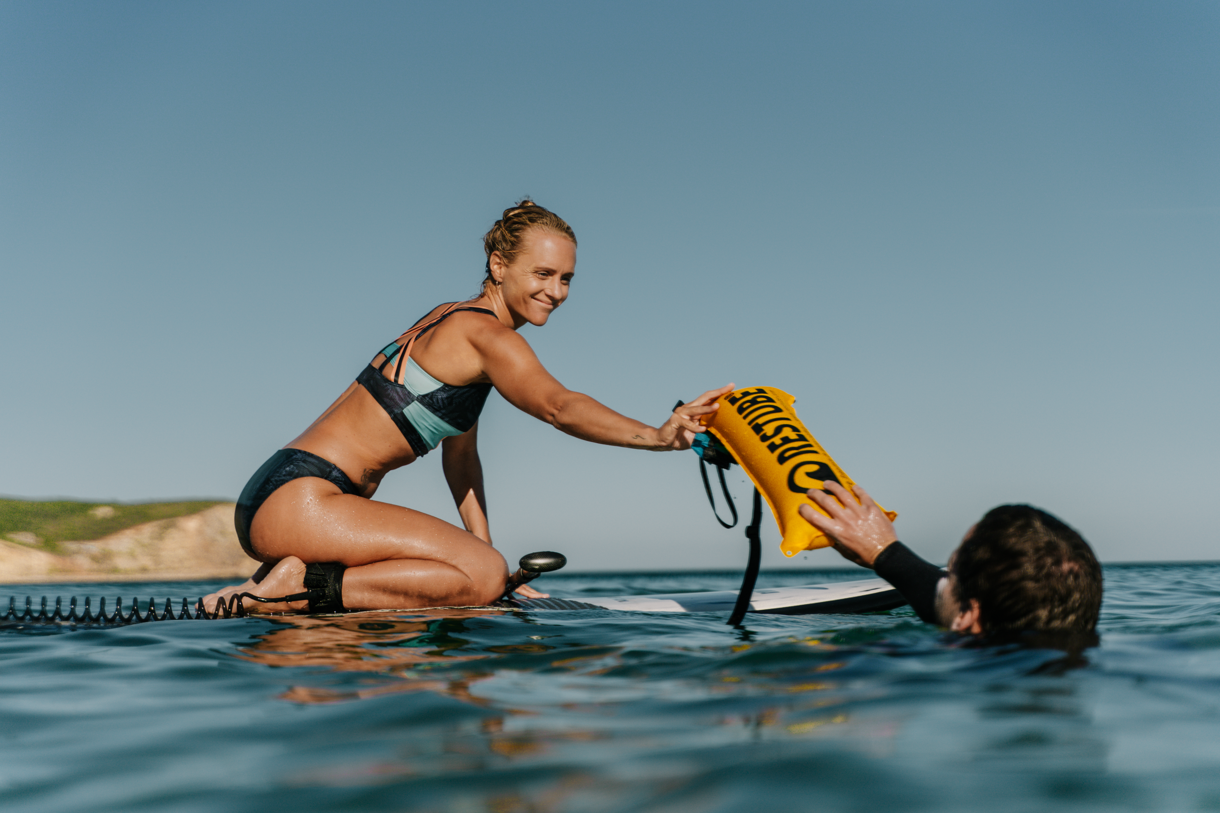 Woman on paddleboard handing RESTUBE buoy to swimmer in the water.