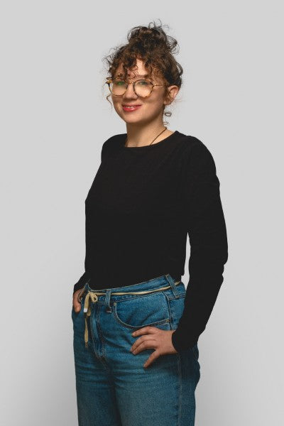 Full-body shot: Curly-haired woman (Sandra Jones) with round glasses, black long-sleeve shirt, and jeans – one hand in pocket, confident look against grey studio background.