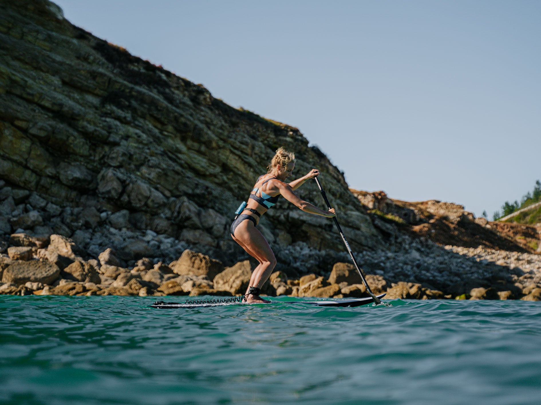 Stand-up paddler with RESTUBE buoy at her back near a rocky coast.