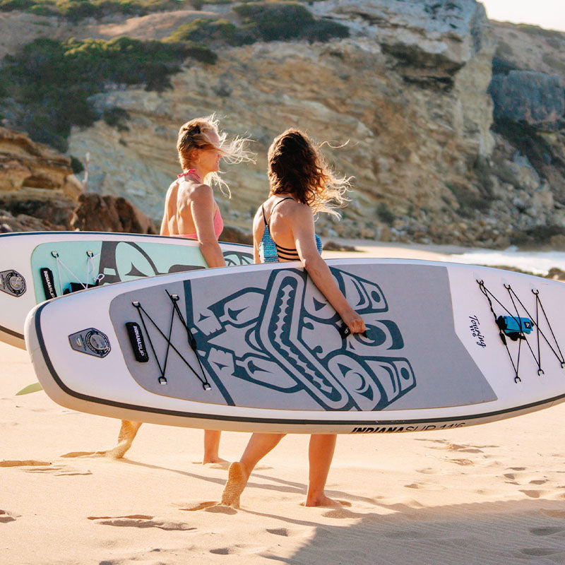 Two women carrying stand-up paddleboards along the beach – ready for water adventures.