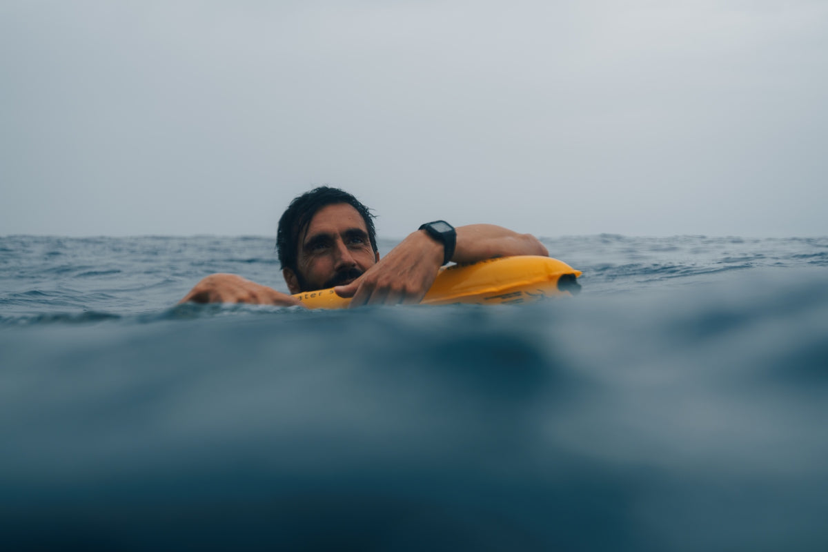 A man floating in open water holding onto a yellow RESTUBE safety buoy, overcast sky in the background