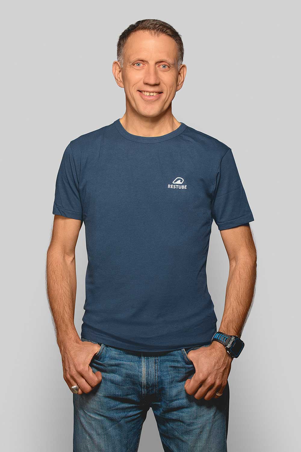 Full-body shot: An Athletic man (Lothar Keppler) in navy RESTUBE T-shirt, hands casually in jeans pockets, smiling in front of neutral grey background.