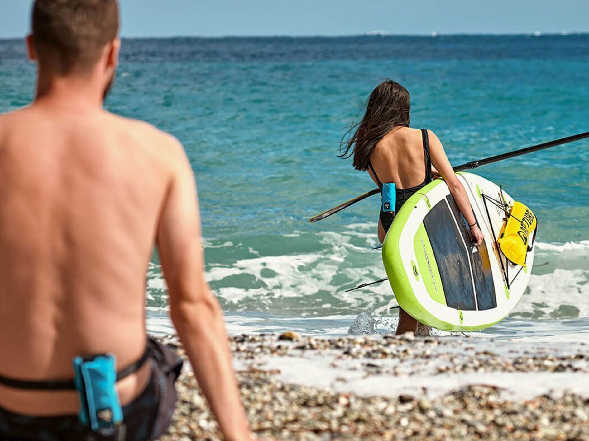 Woman with SUP board and RESTUBE buoy on the beach, carrying the board into the water, man with RESTUBE buoy in the foreground, sitting on the ground.