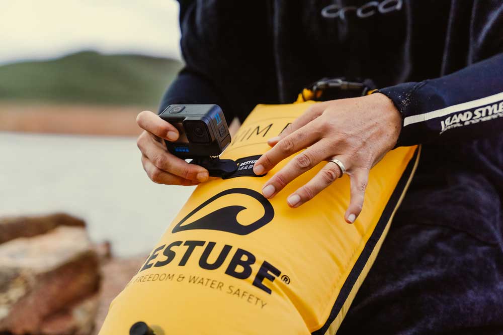 Person attaching a camera to a yellow RESTUBE swim buoy by the water.