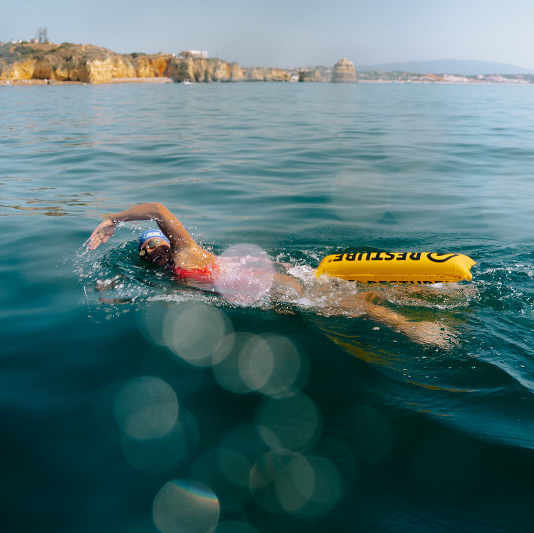 Female swimmer with yellow RESTUBE buoy in open sea near rocky coast.