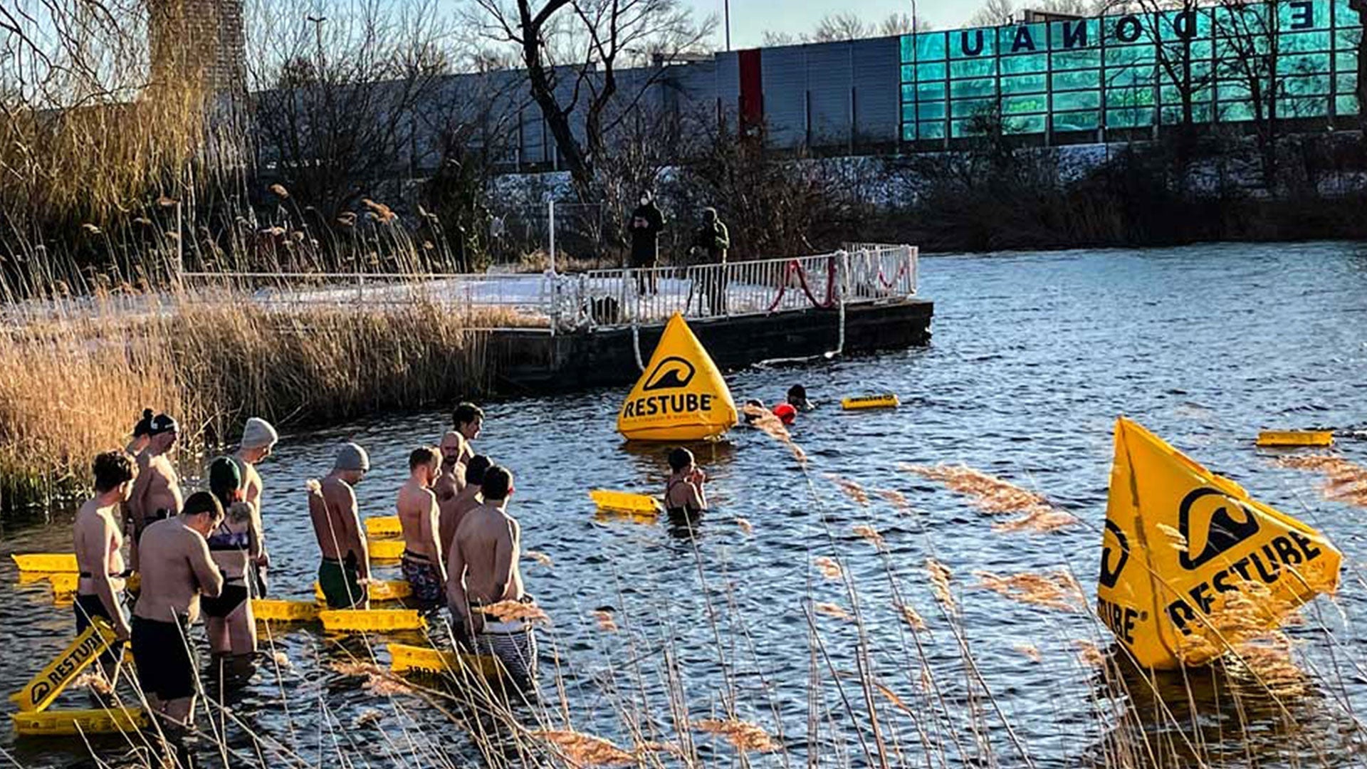 Group of ice swimmers entering cold open water, standing between yellow RESTUBE safety buoys at a marked winter swimming spot.