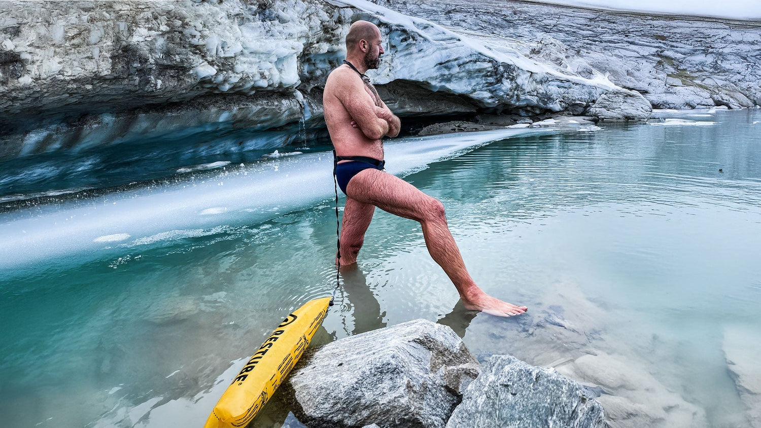 A person wearing swim trunks stands knee-deep in icy glacial water, arms crossed, next to a yellow Restube safety buoy, with frozen rock formations and a turquoise lake in the background.