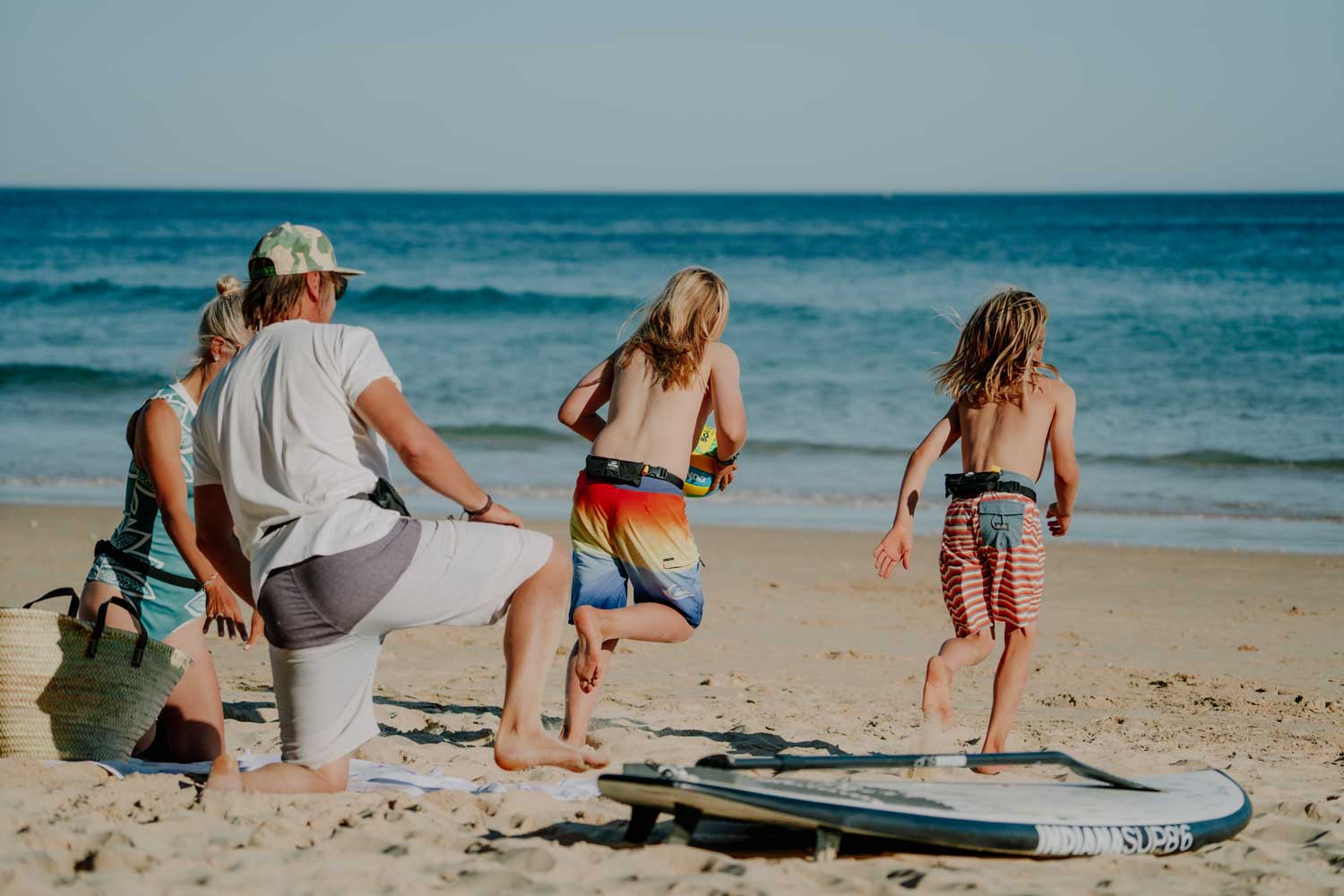 Familie ist am Strand und die zwei Kinder laufen mit einem Ball in Richtung Meer während die Eltern hinterher gucken
